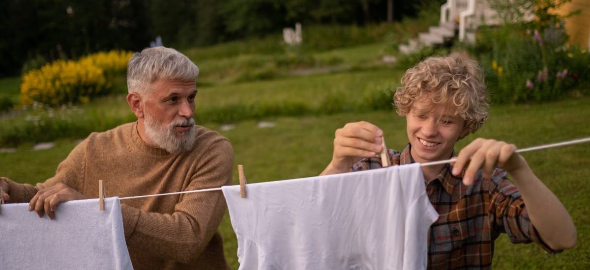 Elderly man and teenage boy joyfully hanging laundry in a sunny garden setting.