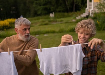 Elderly man and teenage boy joyfully hanging laundry in a sunny garden setting.