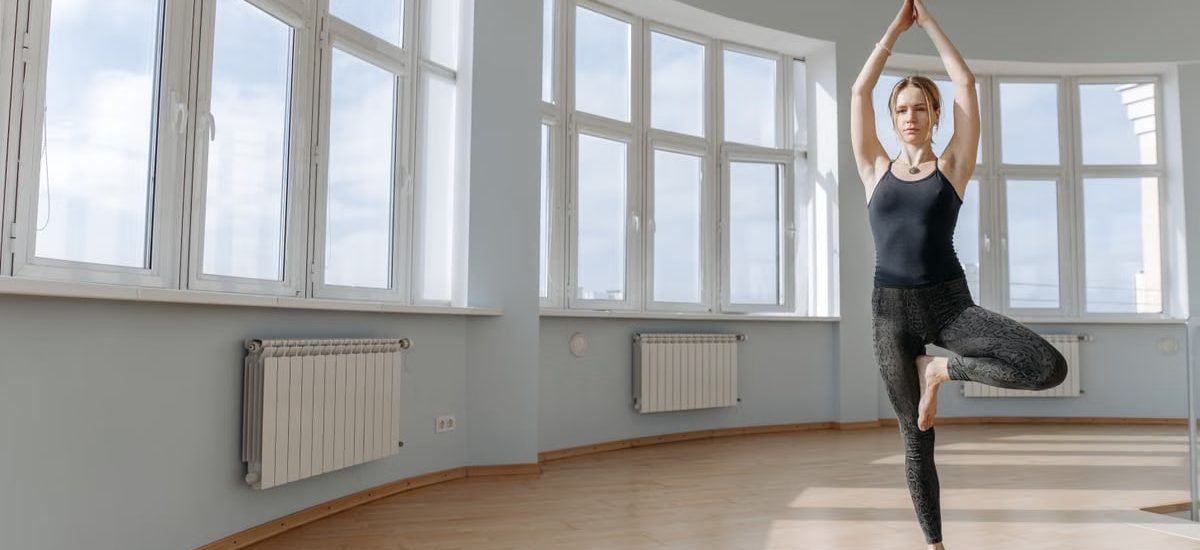A woman performs a yoga pose in a spacious room with large windows, highlighting fitness and wellness.